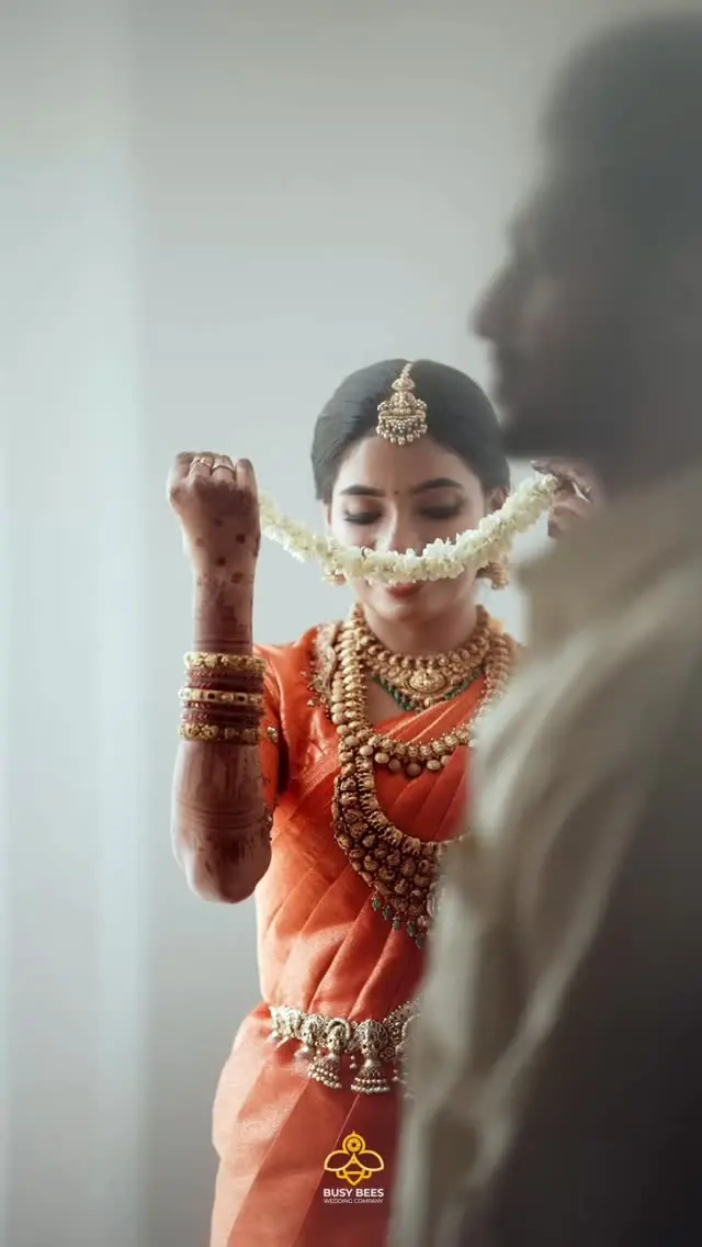 Abhijith & Chinnu: bride lifts a jasmine garland, henna across her hands, in an orange kasavu saree