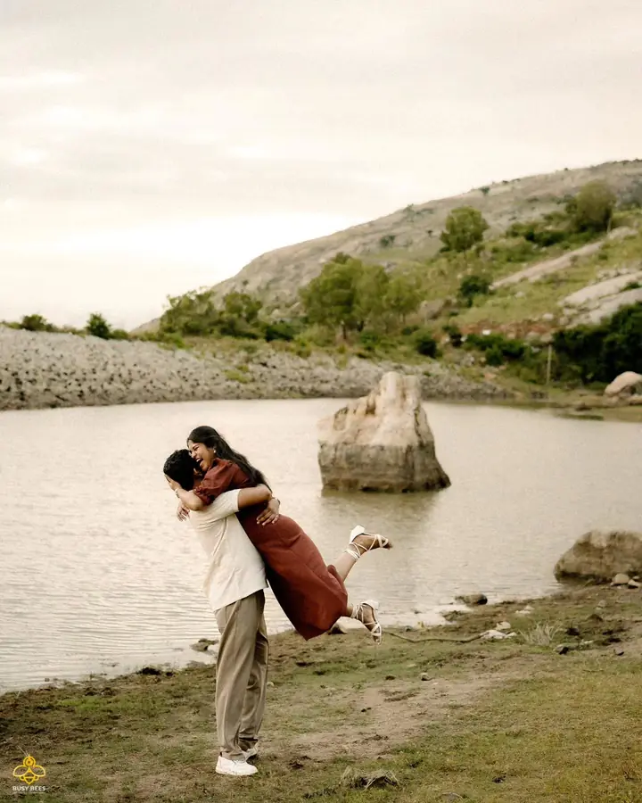 Couple embrace beside a still lake with hills rising behind them