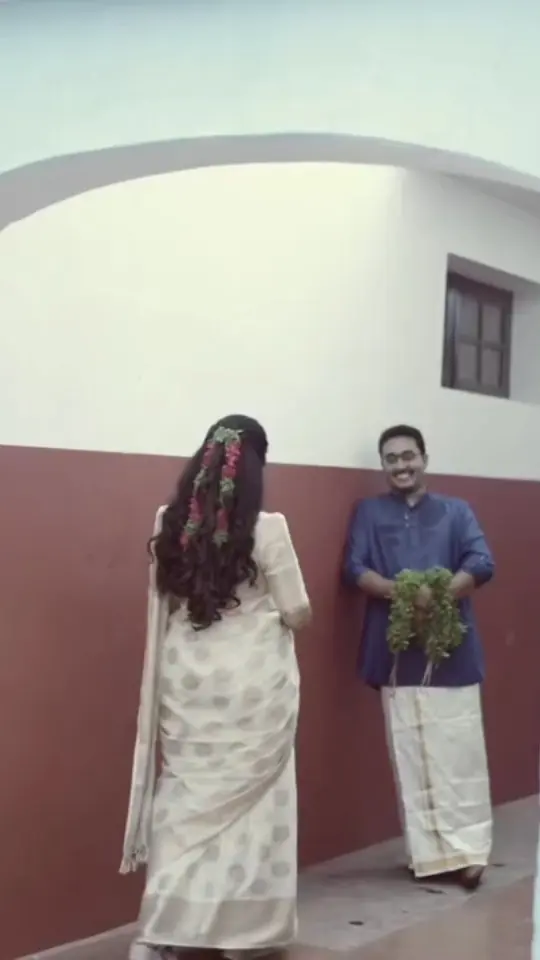 Bride in a white kasavu saree turns towards the groom against an ochre wall
