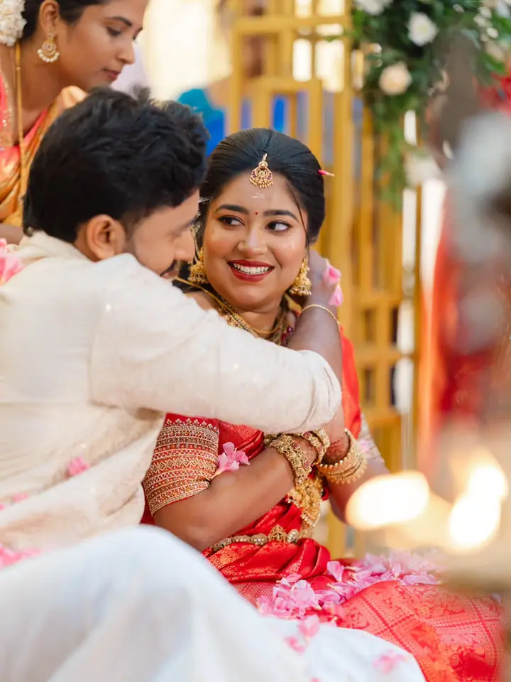 The groom holds the bride from behind, both laughing, amid lamps and flowers