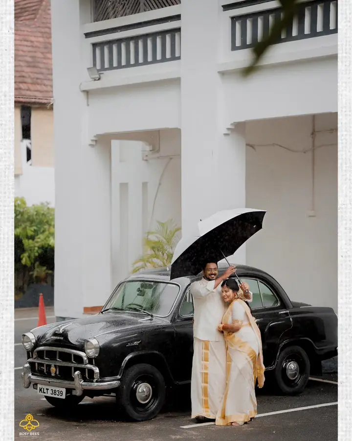 Couple shelter under one umbrella beside a vintage black Ambassador car